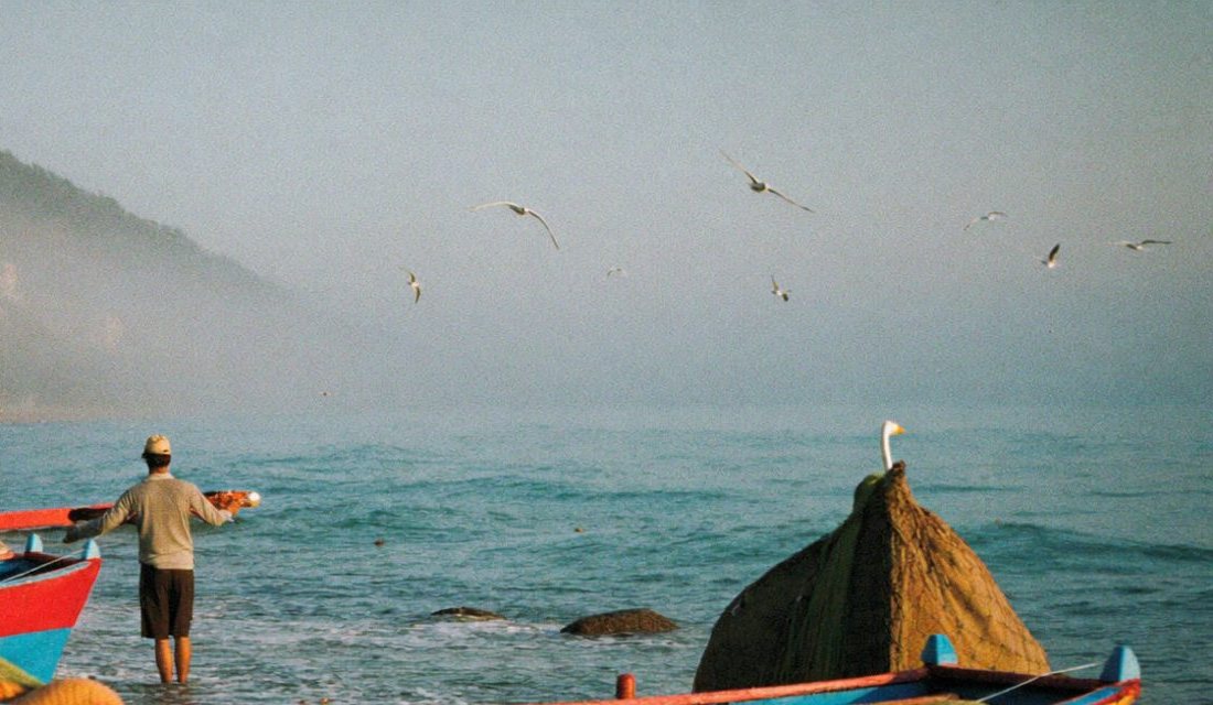 Una caleta de pescadores al amanecer: botes de colores sobre la playa de piedras y un pescador mirando el mar.