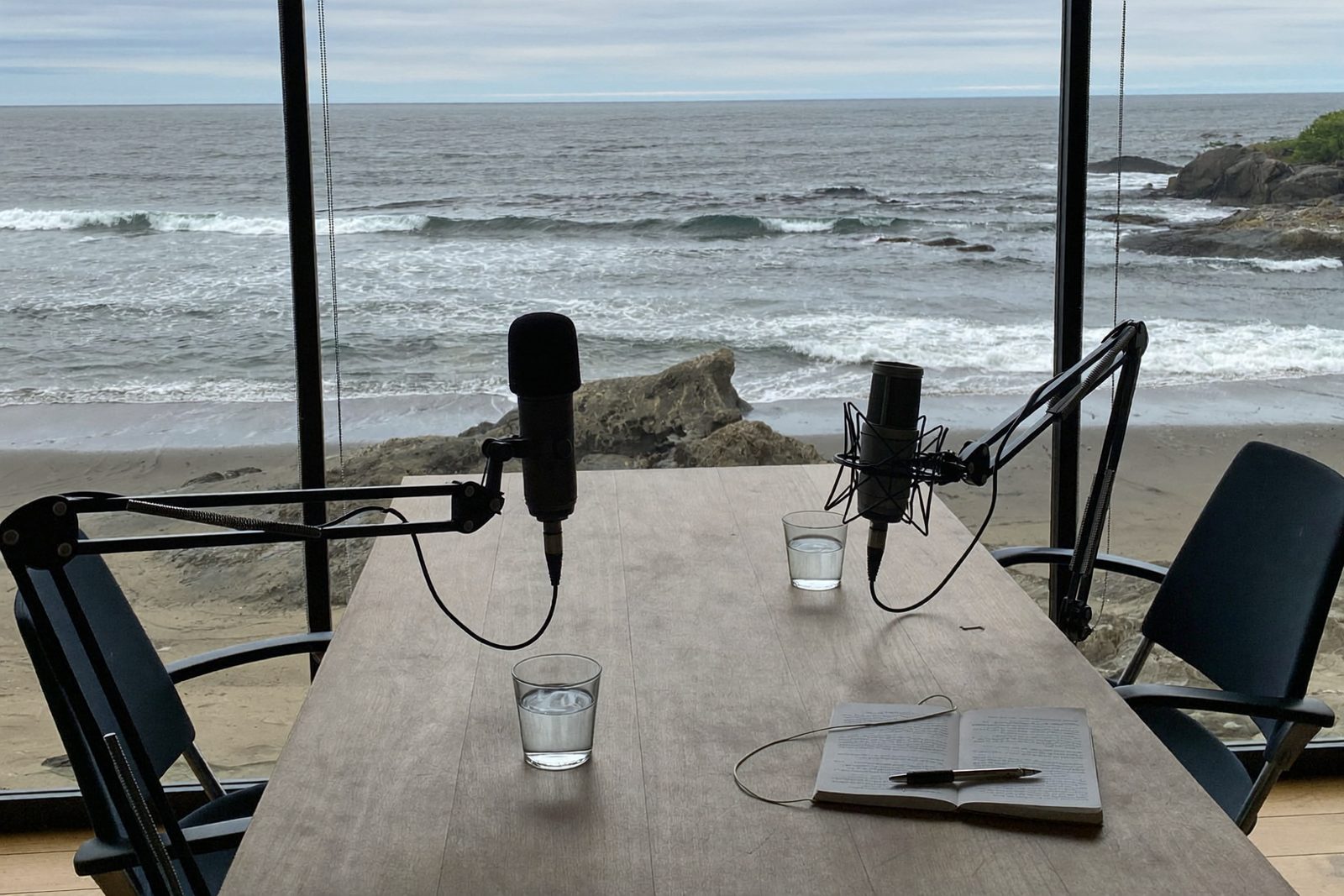 Dos micrófonos sobre una mesa de madera frente a un ventanal que da a una costa rocosa con oleaje.